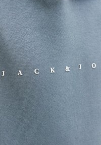 Gaiši zilas audums ar gludu faktūru, uz kura horizontāli pa vidu ir uzraksts "JACK & J" baltos, izceltiem burtiem.