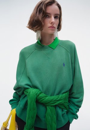 Kvinne med kort brunt hår har på seg en overdimensjonert grønn genser, grønn kabelstrikket genser bundet i midjen, store hvite øredobber og holder en gul håndveske.