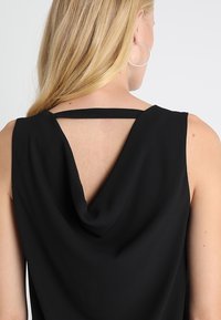 Haut noir sans manches avec un dos drapé en col bénitier et une fine bretelle traversant le haut du dos, texture lisse et silhouette élégante.