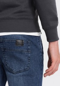 Sudadera gris oscura con puños acanalados, llevada sobre una camisa blanca, combinada con jeans de mezclilla azul que presentan un parche de cuero en el bolsillo trasero.