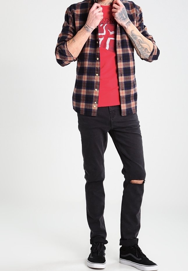 Homme portant un t-shirt graphique rouge sous une chemise à carreaux, un jean noir déchiré et des baskets noires, ajustant le col de sa chemise.