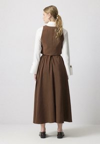 Vestido largo marrón con corpete ajustado y falda fruncida, combinado con una camisa blanca con cuello. Presenta detalles de botones en las mangas.