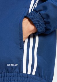 Marinejakke med elastisk manchet og lomme, med hvide tre-stribe detaljer og et Aeroready-logo nær kanten. Glat tekstur.