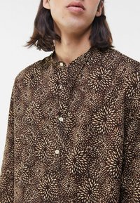 Brun långärmad skjorta med ett detaljerat geometriskt mönster i krämvitt, med en liten krage och knappslå vid halsringningen.