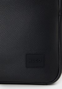 Black textured leather bag featuring a raised logo patch with the name "HUGO" embossed. Smooth edges form a rectangular shape.