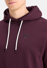 Burgunderfarbener Hoodie mit großer Kapuze, weißen Kordeln und einem kleinen quadratischen Logo-Patch auf der Brust. Weicher, strukturierter Stoff.