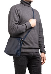 Borsa a tracolla in pelle blu navy con finitura liscia, forma rettangolare e tracolla regolabile, tenuta da una persona che indossa un dolcevita grigio.