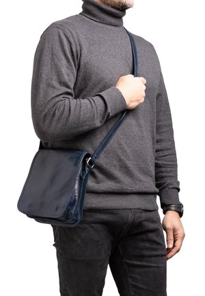 Borsa a tracolla in pelle blu navy con finitura liscia, forma rettangolare e tracolla regolabile, tenuta da una persona che indossa un dolcevita grigio.