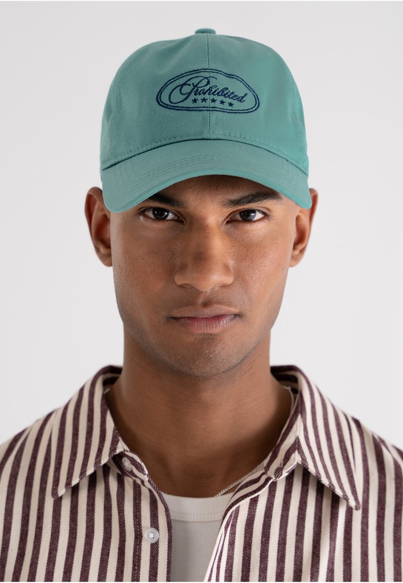 Joven con gorra verde con texto "Chabilited" y estrellas, camisa de rayas abotonada sobre camiseta blanca, fondo neutro.