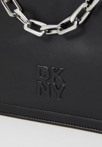 Melna ādas rokassoma ar teksturētu virsmu, iegrieztu "DK NY" logo un sudraba ķēdes akcentu, ar gludu vīli malā.