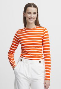 Långärmad topp i orange och vita ränder, tillverkad av elastiskt tyg, med rund halsringning. Kombinerad med högmidjade vita byxor med knapppdetaljer.