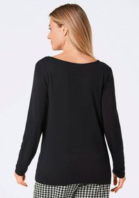 Schwarzes Langarmshirt mit weitem Ausschnitt, aus glattem Stoff gefertigt. Das Modell trägt gemusterte Hosen mit einem Hahnentrittmuster.