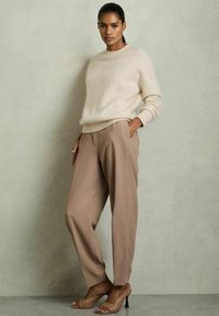 Beige kevyt neulepaita pyöreällä kaula-aukolla ja pitkillä hihoilla, yhdistettynä korkeavyötäröisiin taupe-värisiin muotihousuihin ja ihonvärisiin avokkaisiin.