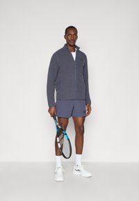 Pelēka jaku ar rāvējslēdzēju, ar sānu kabatām, uz muguras uzvilkta virs baltas krekliņa, valkājot tumši zilu šortu. Rokā tur tenisa raketi, uz kājām ir baltas kedas.