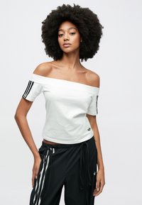 Fehér, vállra húzható pamut póló, rövid ujjakkal, fekete díszcsíkokkal. Kis fekete logó a szegélyen. Fekete melegítőnadrággal párosítva.