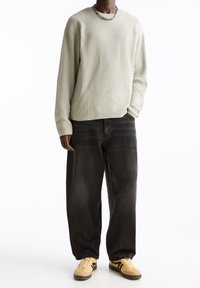 Hellgrauer gerippter Pullover mit langen Ärmeln, kombiniert mit locker sitzenden schwarzen Jeans und beigen Sneakern mit schwarzen Akzenten. Schlichter Perlenhalsreifen getragen.