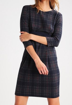 Femme portant une robe à carreaux foncés longueur genou avec des manches trois-quarts, debout les bras croisés sur le ventre.