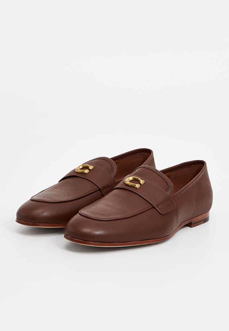Mocassins en cuir marron avec une semelle plate, présentant un détail en métal doré proéminent à l'avant. Texture lisse et design classique.