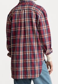 Rückenansicht einer Person, die ein kariertes Hemd in Rot, Marineblau und Creme mit hochgekrempelten Ärmeln über einem weißen Langarmshirt und blauen Jeans trägt.