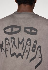 Szara bawełniana bluza z dużą czarną grafiką oczu oraz słowem "KARMA" z gestami dłoni na plecach. Prosty, casualowy design.