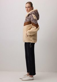 Sudadera beige difusa con capucha de leopardo y panel marrón, combinada con jeans negros y zapatillas blancas, mostrando un ajuste relajado.