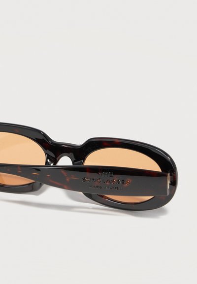 Ρετρό οβάλ γυαλιά ηλίου με καφετί φακούς, διπλωμένα με λογότυπο και το κείμενο "Κατασκευασμένα στην Ιταλία" στο μπράτσο.