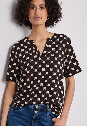 Frau mit lockigem dunklem Haar, die goldene Creolen, eine kurzärmelige Bluse mit schwarz-weißem geometrischem Muster und blaue Jeans trägt.