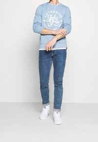 Sudadera azul claro con estampado de logo blanco, combinada con jeans ajustados azules y zapatillas blancas. Diseño casual y cómodo.