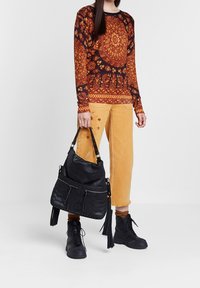Bolso de cuero negro con superficie texturizada y bolsillos con cremallera, complementado por una parte superior mostaza con patrones intrincados y pantalones de corte relajado.