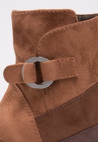 Bottine en suède brun avec un bout arrondi, des détails cousus et une sangle avec une boucle circulaire en métal. Texture douce.