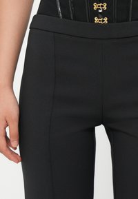 Close-up of a person wearing black fitted pants and a black top with gold hook-and-eye fasteners, showing their left hand relaxed by their side.