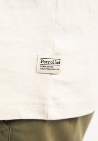 Lichtbeige t-shirt met een rechthoekig merklabel met zwarte tekst. De stof lijkt zacht met een gladde textuur; de zoom is gestikt.