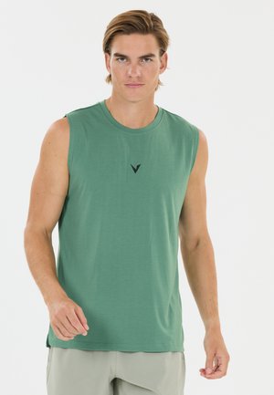 Junger Mann mit hellbraunen Haaren trägt ein ärmelloses grünes Shirt und hellgraue Shorts vor einem schlichten weißen Hintergrund.