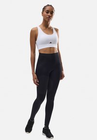 Mallas negras de cintura alta combinadas con un sujetador deportivo blanco con un detalle de recorte. Modelo que lleva zapatillas deportivas negras. Textura suave.