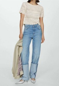 Kremowy krótki top ze falbankami na dole, w połączeniu z jasnoniebieskimi, szerokimi dżinsami. Modelka trzyma beżową kurtkę. Białe czółenka w szpic.