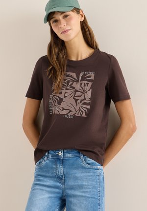 Femme portant un t-shirt marron foncé avec un motif abstrait de feuilles et un texte en français, associé à un jean bleu clair et une casquette verte.
