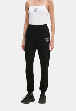 Top in raso bianco con spalline sottili e pantaloni jogger neri con un logo sull'anca sinistra, abbinati a scarpe da ginnastica nere.
