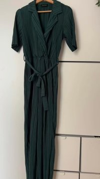 Temno zelena, nabrita kombinacija z kratkimi rokavi, z izrezanim ovratnikom in pasOM iz blaga. Gladka tekstura in dolžina do tal. Ovise na lesenem obešalniku.