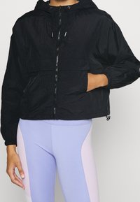 Svart kort jacka med huva, dragkedjestängning och sidofickor. Tillverkad av lätt material, med elastiska cuffs och en avslappnad passform.