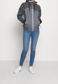 Veste à capuche avec un corps gris, motif à pois sur les manches, et une fermeture éclair blanche. Associée à un jean bleu et des baskets blanches.