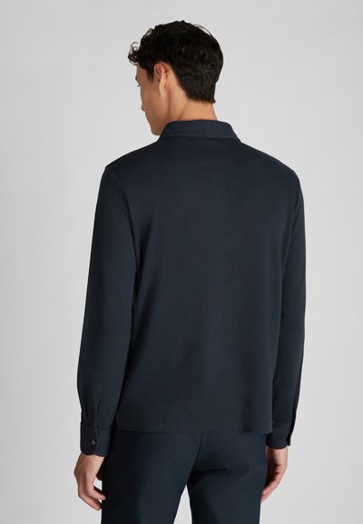 Homme de dos portant une chemise à manches longues bleu marine avec des poignets boutonnés et un pantalon sombre, devant un fond clair.