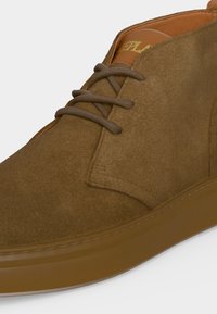 Chaussure en daim marron à lacets avec une semelle marron lisse et une languette en cuir fauve ornée d’inscriptions dorées partielles.