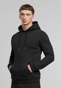 Felpa nera con cappuccio realizzata in morbido misto cotone. Presenta una tasca frontale, un cordino regolabile e polsini e orlo a costine. Vestibilità classica.