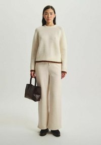 Pull en tricot beige, pantalon large crème, et un petit sac fourre-tout marron foncé. Un tissu texturé et des poignets contrastants décorent la tenue.