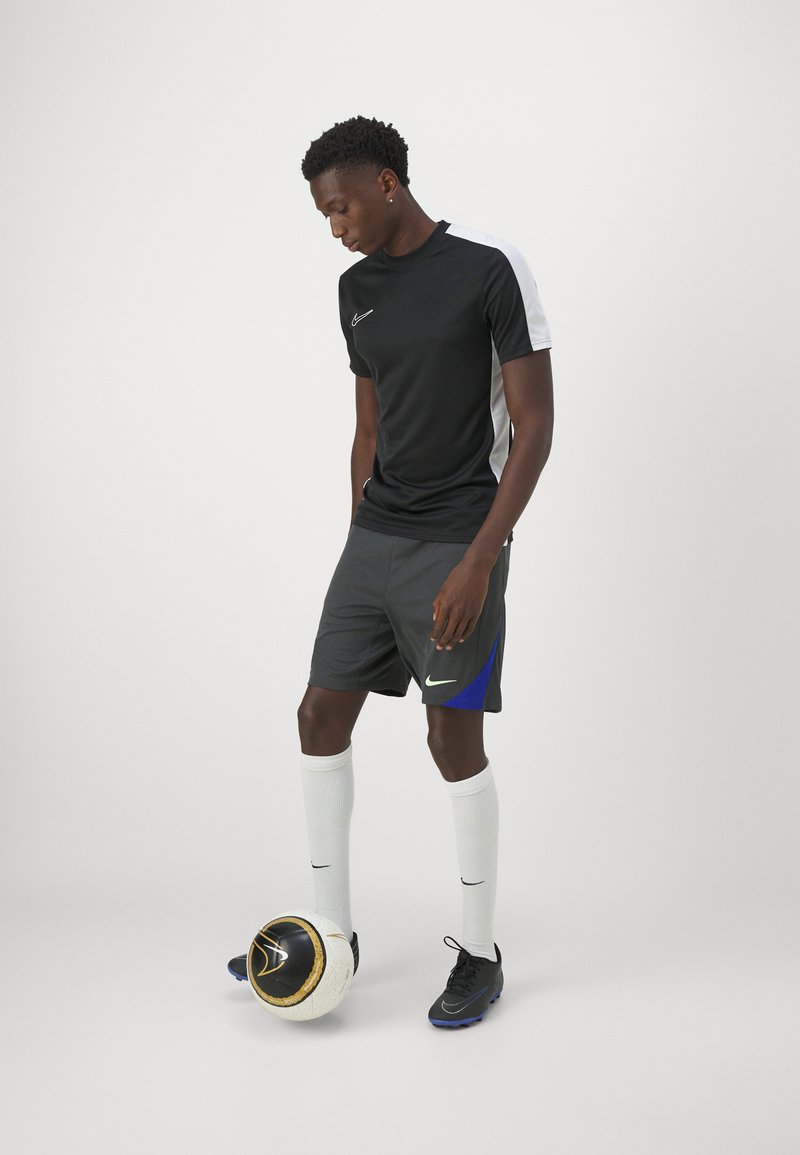 Camiseta atlética negra con acentos blancos, pantalones cortos grises con detalles en azul, calcetines blancos y zapatos de fútbol negros, de pie al lado de un balón de fútbol con un diseño.