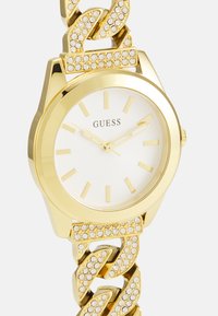 Orologio dorato con quadrante bianco, dotato di indicatori delle ore e di un bracciale a maglie arricchito da cristalli trasparenti e una finitura lucida.