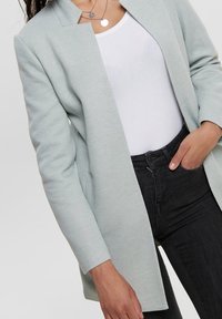 Blazer largo de color verde claro y textura, con solapa, frontal abierto y mangas largas, combinado con una blusa blanca y jeans negros.