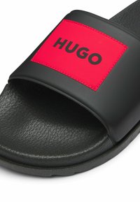Musta kuminen liukusandaali, jossa on leveä punainen hihna, jossa on kohokuvioitu musta "HUGO" -logo, teksturoidut mustat pohjat ja kestävä musta pohja.