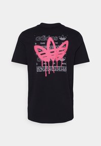 Svart t-shirt med vit Adidas-text och logotyp överlagrad med stort rosa graffiti-stil treflor-mönster som droppar färg.