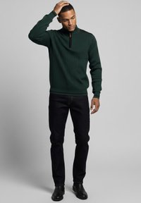 Jersey verde acanalado con cuello de cremallera, detalles negros en los hombros, combinado con vaqueros oscuros y zapatos negros. Diseño simple y ajustado.